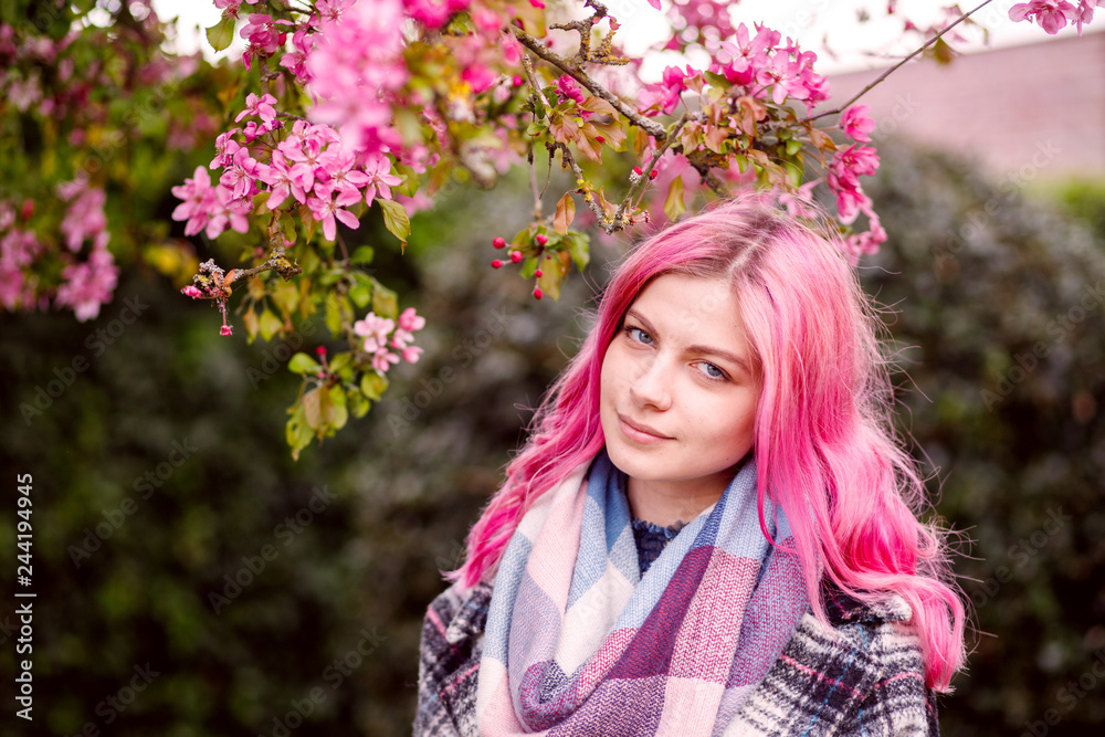 Fototapeta premium young beautiful girl with pink hair standing near a tree with flowers, pink flowers, spring, sun, happiness