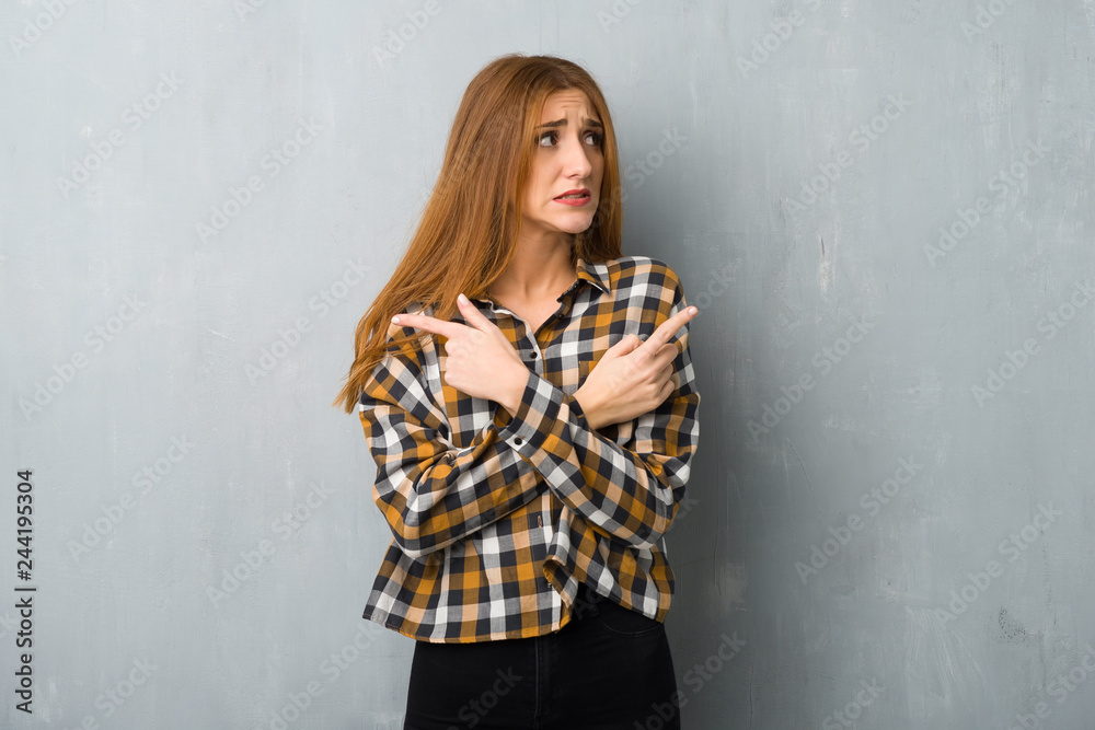Young redhead girl over grunge wall pointing to the laterals having doubts Young redhead girl over grunge wall pointing to the laterals having doubts