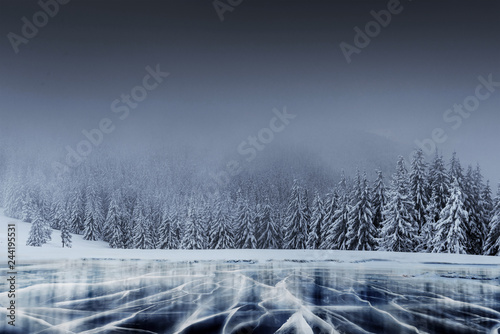 Fototapeta Naklejka Na Ścianę i Meble -  Majestic winter landscape with cracks on the surface of the blue ice. Frozen lake in winter mountains. A dramatic scene with low black clouds, a calm before the storm