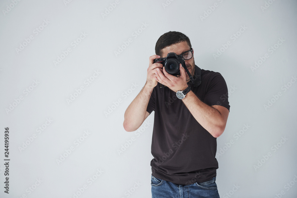 A young hipster man in eyepieces holds a DSLR camera in hands standing against a white wall background A young hipster man in eyepieces holds a DSLR camera in hands standing against a white wall background