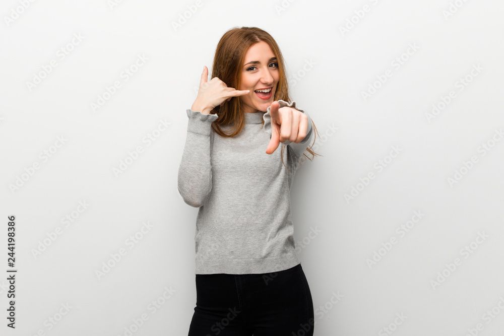 Redhead girl over white wall making phone gesture and pointing front Redhead girl over white wall making phone gesture and pointing front