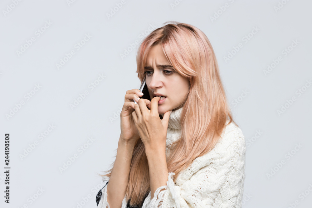 Studio portrait of blonde girl in white sweater talking on the phone, said terrible news, sad look, gnawing nails/ found out what happened, bad news, nervous feeling,experiences of fear/people concept Studio portrait of blonde girl in white sweater talking on the phone, said terrible news, sad look, gnawing nails/ found out what happened, bad news, nervous feeling,experiences of fear/people concept