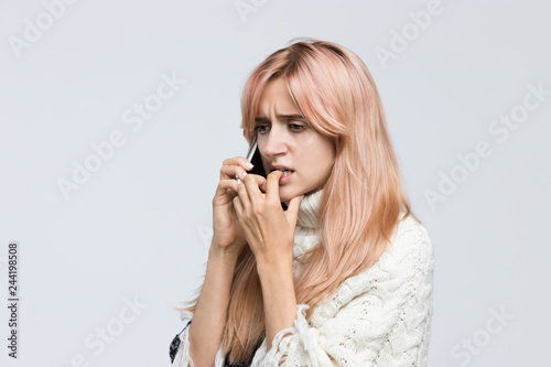 Studio portrait of blonde girl in white sweater talking on the phone, said terrible news, sad look, gnawing nails/ found out what happened, bad news, nervous feeling,experiences of fear/people concept