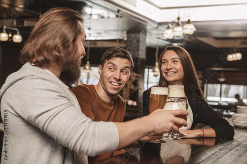 Handsome young man smiling happily, enjoying resting at the bar with his friends. Group of friends having drinks at the local beer pub. Two men and beautiful woman drinking beer. Celebration concept