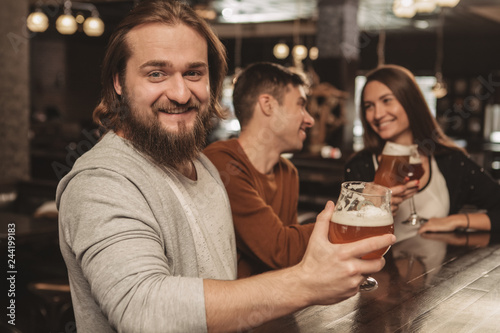 Bearded happy man smiling to the camera joyfully with a glass of beer in his hand, his friends chatting on the background. Cheerful man enjoying beer party with his friends. Leisure, recreation concep