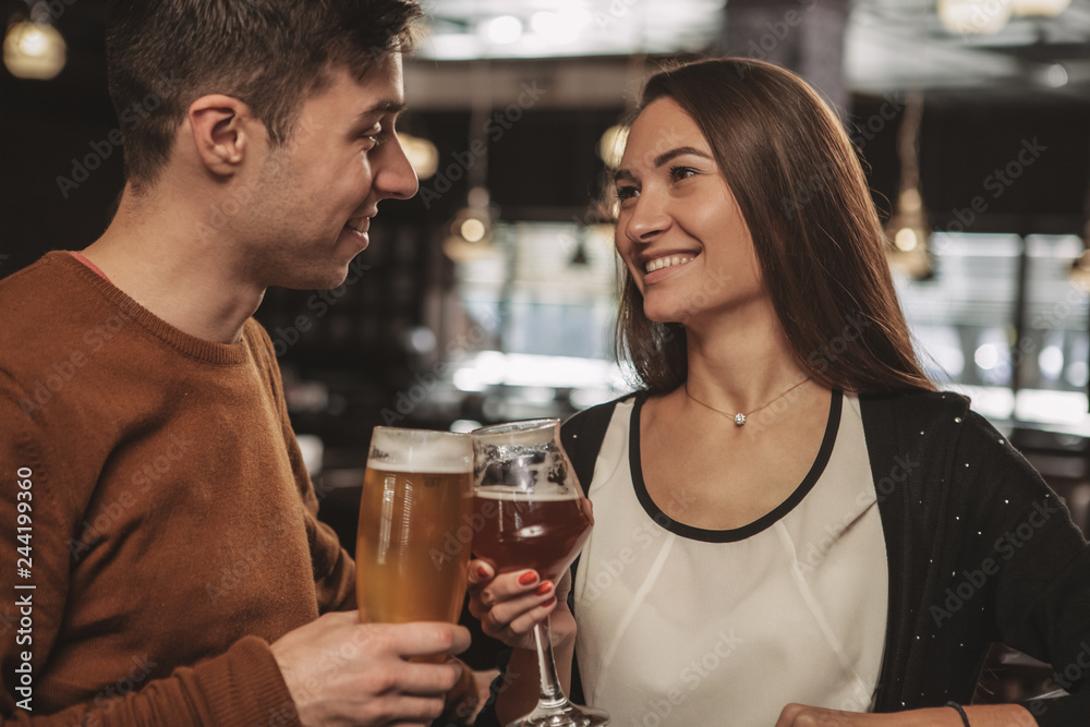 Beautiful happy couple looking at each other with love, celebrating their anniversary, having drinks at the restaurant together. Beautiful woman and her handsome boyfriend drinking craft beer
