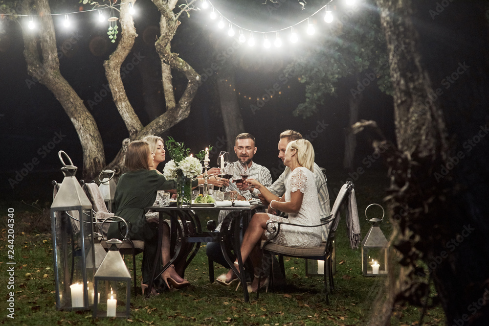 Beautiful garlands create a sense of fairy tale. Group of adult friends have a rest and conversation in the backyard of restaurant at evening time - obrazy, fototapety, plakaty 