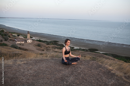 young woman Practicing Yoga in Lotus pose sitting on the edge of rock against of see and lighthouse. Padmasana
