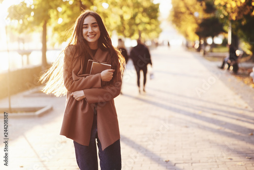 Front view of young, smiling, pretty brunette girl standing and holding small stack of books in her hands, wearing casual coat at autumn sunny park alley on blurry background with a copy space