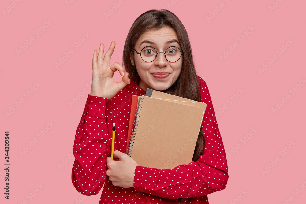 Brunette young college student demonstrates everything is alright, shows okay gesture, holds notepads and pencil, confident in successfully passed exam, isolated over pink wall. Ok, I agree with you Brunette young college student demonstrates everything is alright, shows okay gesture, holds notepads and pencil, confident in successfully passed exam, isolated over pink wall. Ok, I agree with you