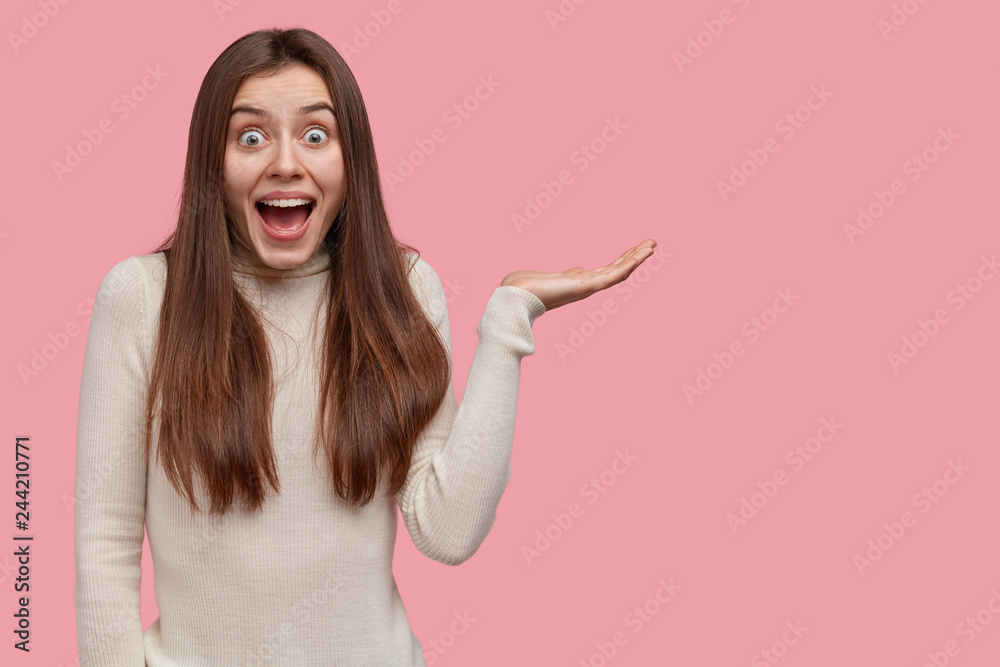 Studio shot of happy dark haired woman raises palm, feels excited, holds copy space, dressed casually, demonstrates something, satisfied to show something awesome and cool, isolated on pink wall Studio shot of happy dark haired woman raises palm, feels excited, holds copy space, dressed casually, demonstrates something, satisfied to show something awesome and cool, isolated on pink wall