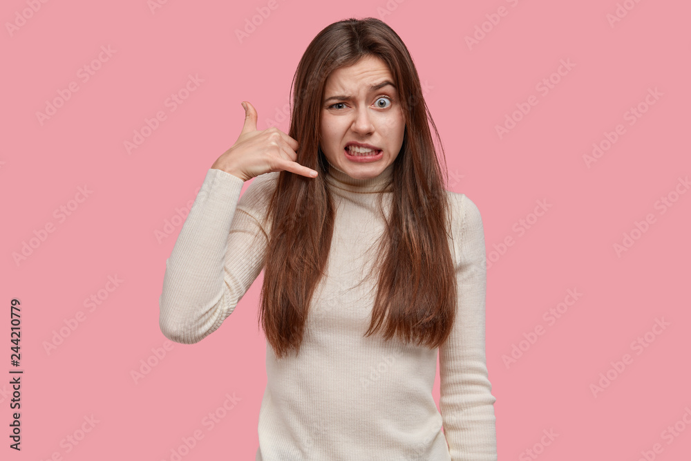 Call me back sign. Frustrated dark haired woman makes phone gesture, raises eyebrows, wears in white clothes, isolated over pink background. Communication, symbols and body language concept. Call me back sign. Frustrated dark haired woman makes phone gesture, raises eyebrows, wears in white clothes, isolated over pink background. Communication, symbols and body language concept.