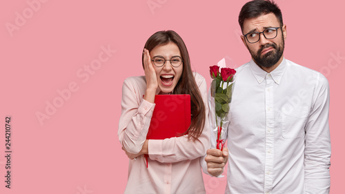 Overjoyed glad woman has first date, expresses positive emotions, awkward guy stands near with bouquet of roses, wears white shirt, doesnt know how to behave. Romantic couple poses over pink wall