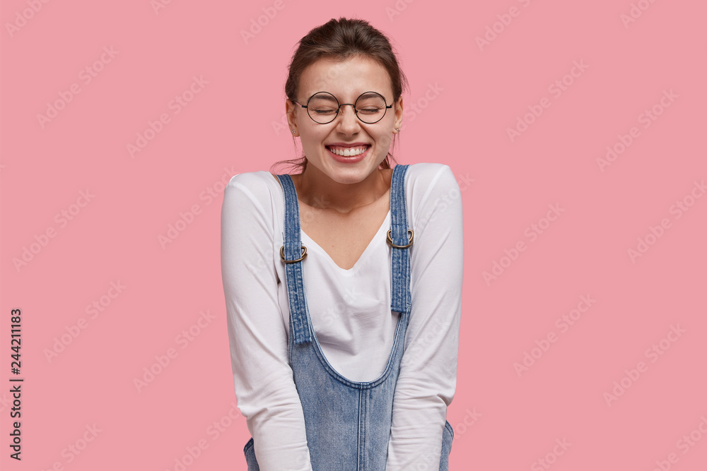 People, satifaction and joy concept. Optimistic glad woman closes eyes with pleasure, smiles broadly, wears casual clothes, expresses good emotions, isolated over pink background laughs at funny thing People, satifaction and joy concept. Optimistic glad woman closes eyes with pleasure, smiles broadly, wears casual clothes, expresses good emotions, isolated over pink background laughs at funny thing