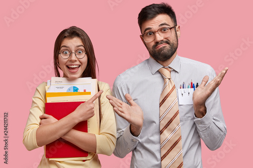 Pretty positive woman points at hesitant guy who spreads palms in bewildermet, tells to ask him this question, carries necessary documentation, stand next to each other, isolated over pink backgound