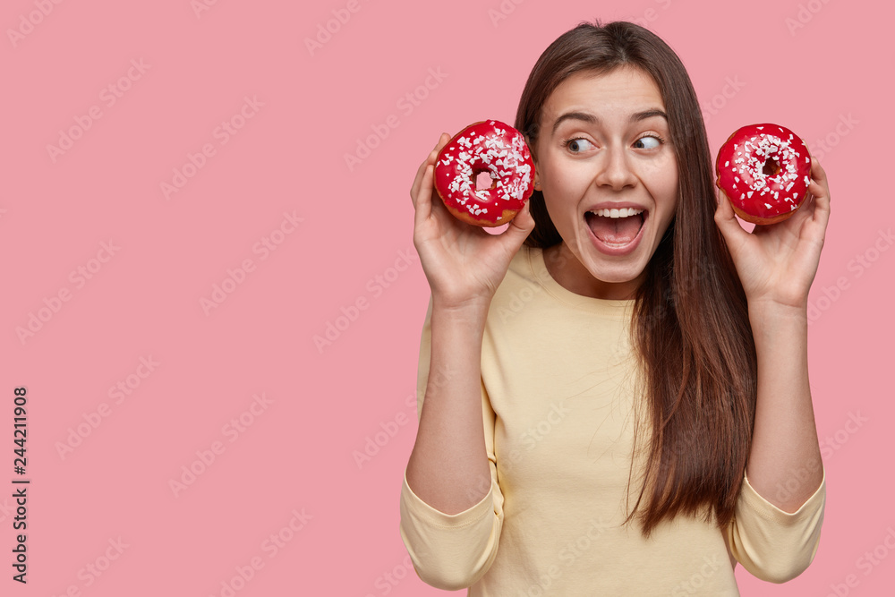 Joyful youngster with dark hair, carries two delicious glazed donuts, opens mouth widely, dressed in casual clothes, isolated over pink background with empty space for your advertisement or text Joyful youngster with dark hair, carries two delicious glazed donuts, opens mouth widely, dressed in casual clothes, isolated over pink background with empty space for your advertisement or text