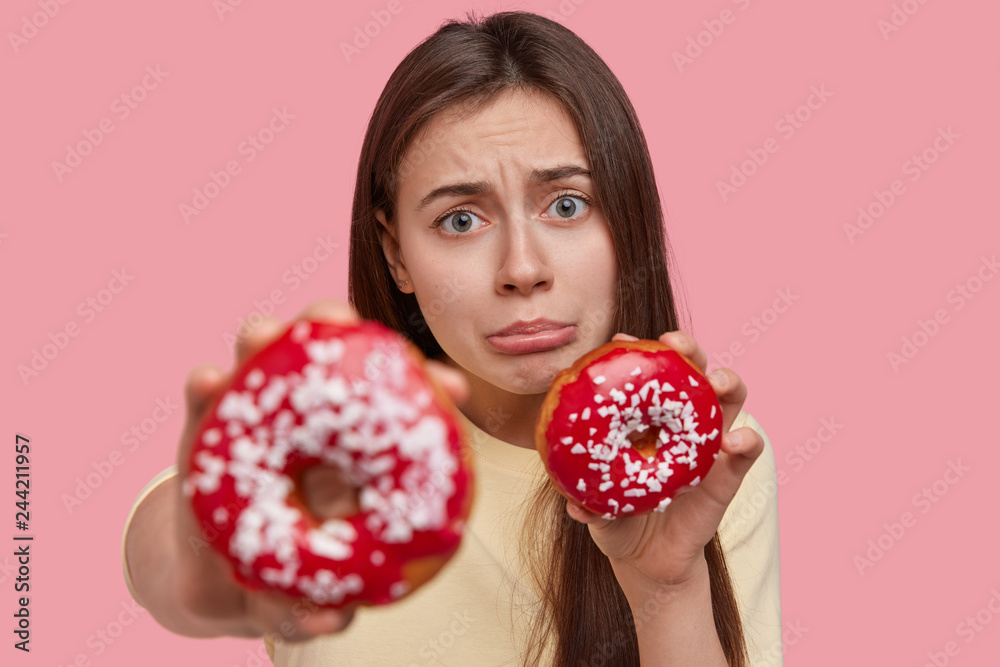 Upset brunette lady pures lips, feels dissatisfied, cant eats what she wants, keeps to diet, carries red doughnuts, isolated over pink background. Hungry woman feels temptation, holds junk food Upset brunette lady pures lips, feels dissatisfied, cant eats what she wants, keeps to diet, carries red doughnuts, isolated over pink background. Hungry woman feels temptation, holds junk food