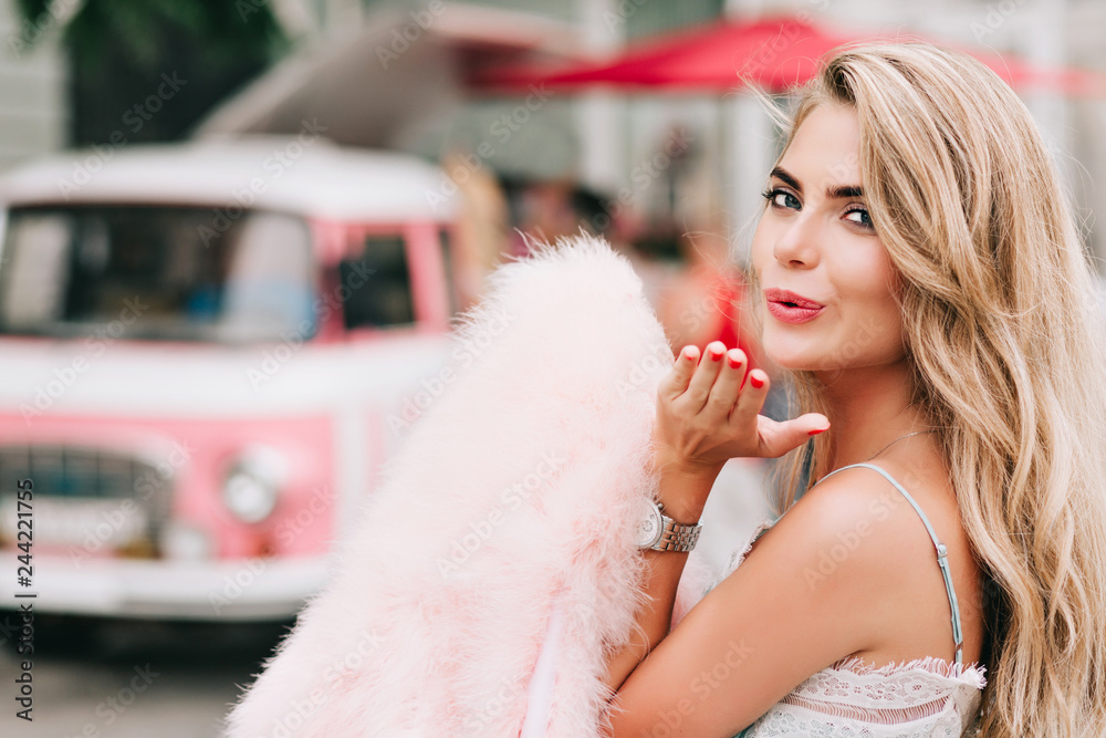 Closeup portrait attractive girl holding pink fur stole in hand on retro car background. She has long blonde hair, bare shoulder, sending a kiss to camera Closeup portrait attractive girl holding pink fur stole in hand on retro car background. She has long blonde hair, bare shoulder, sending a kiss to camera