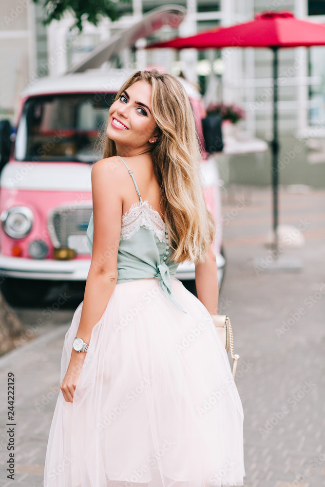 View from back pin up styled girl with long blonde hair walking on street. She wears light tulle skirt, has bare shoulder, smiling to camera View from back pin up styled girl with long blonde hair walking on street. She wears light tulle skirt, has bare shoulder, smiling to camera