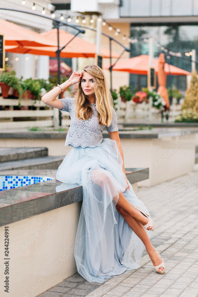 Attractive girl with long blonde hair in blue long tulle skirt sitting on terrace background. She keeps hand on head and looking to camera