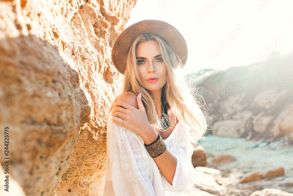 Portrait of attractive blonde girl with long hair posing to the camera on beach on rock background. She wears white shirt, hat, ornamentation.