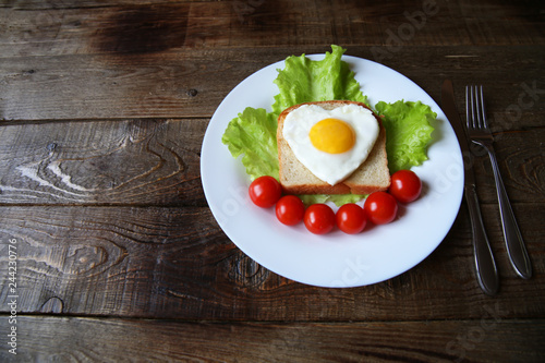 fried eggs with a yellow yolk in the form of a heart on bread, decorated with green salad and red cherry tomatoes on a white plate and wooden rustic background with space for text 