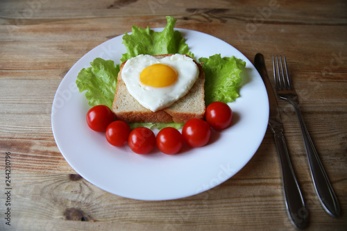 fried eggs with a yellow yolk in the form of a heart on bread, decorated with green salad and red cherry tomatoes on a white plate and wooden rustic background 