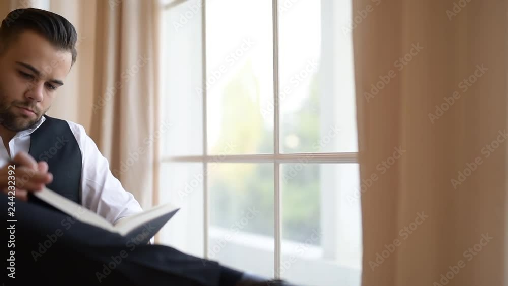 A young man sitting on armchair and flips through a book ove background of window. A young man sitting on armchair and flips through a book ove background of window.