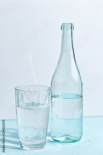 A glass bottle with clear water and a glass on a white-blue background. Minimalistic creative concept.