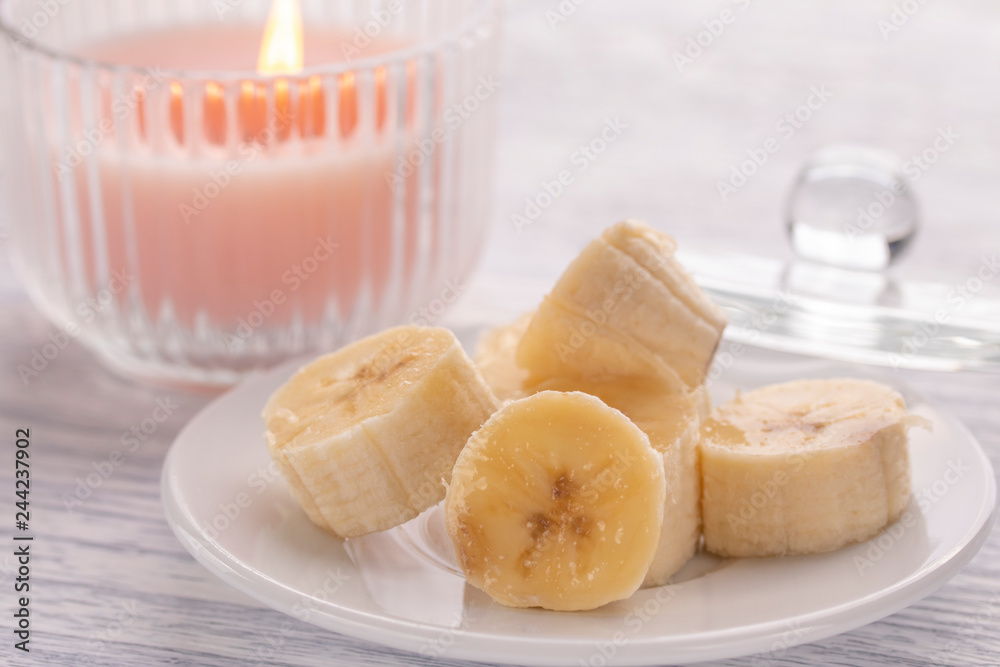 Sliced banana on a white plate and a light wooden table. Pink burning candle nearby. Sliced banana on a white plate and a light wooden table. Pink burning candle nearby.