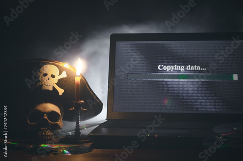 Illegal data copying concept. Cybercrime. Computer piracy background. Pirate hat, human skull, laptop and compact disc on a table.