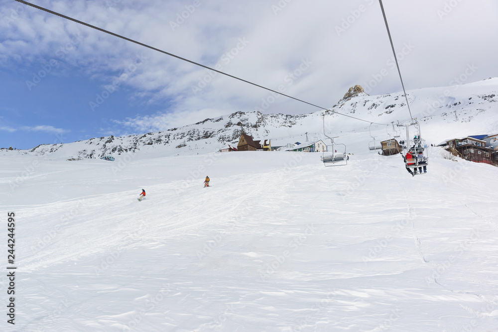 A two-seat cable car and a ski track with snowboarders in the ski resort of Dombay in the middle of the winter season