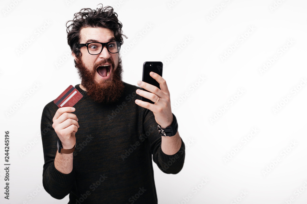 Handsome excited bearde man looking at the screen of mibile and holds his credit card Handsome excited bearde man looking at the screen of mibile and holds his credit card