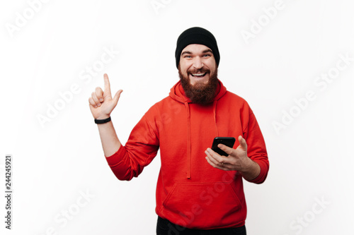 Handsome smiling bearde guy in red pullover is pointing up while hold his mobile and looking at the camera photo