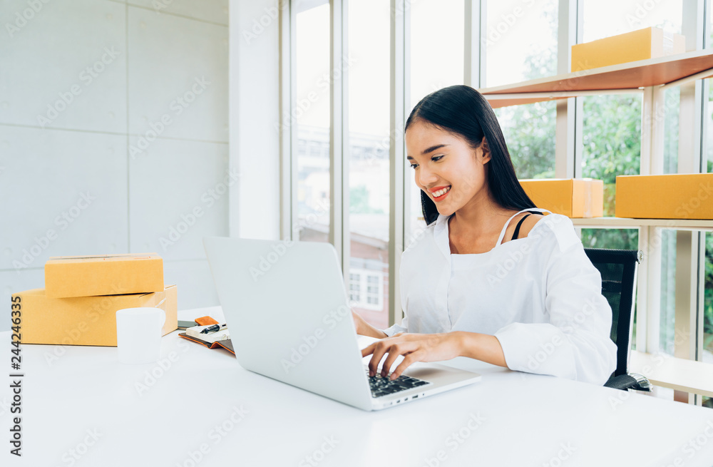 Young Asian casual businesswoman smiling happy using laptop working checking customer's order and products packaging for delivery in modern home office, online shopping business concept
