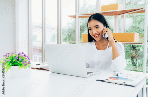 Young asian casual businesswoman using mobile phone and laptop to communicate with customer for order purchasing abd delivering information in modern home office, online shopping business concept photo