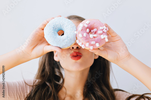 Closeup portrait joyful girl with red lips having fun with colorful donuts against her eyes. Attractive young woman with long hair holding sweets. Good mood, diet concept, brightful colors photo