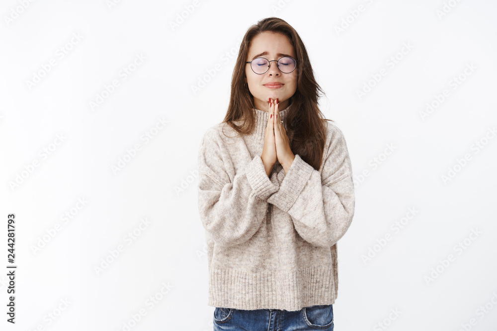 Girl feeling worried and insecure frowning being emotional as close eyes holding hands in pray, making wish praying faithfully with hope things get better, standing nervous and unhappy over gray wall Girl feeling worried and insecure frowning being emotional as close eyes holding hands in pray, making wish praying faithfully with hope things get better, standing nervous and unhappy over gray wall