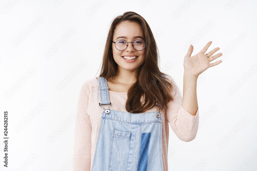 Waist-up shot of cute female newbie waving new coworker hopefully, making friends smiling friendly and happy at camera as raising palm in hello or hi gesture, greeting everyone pleased and joyful