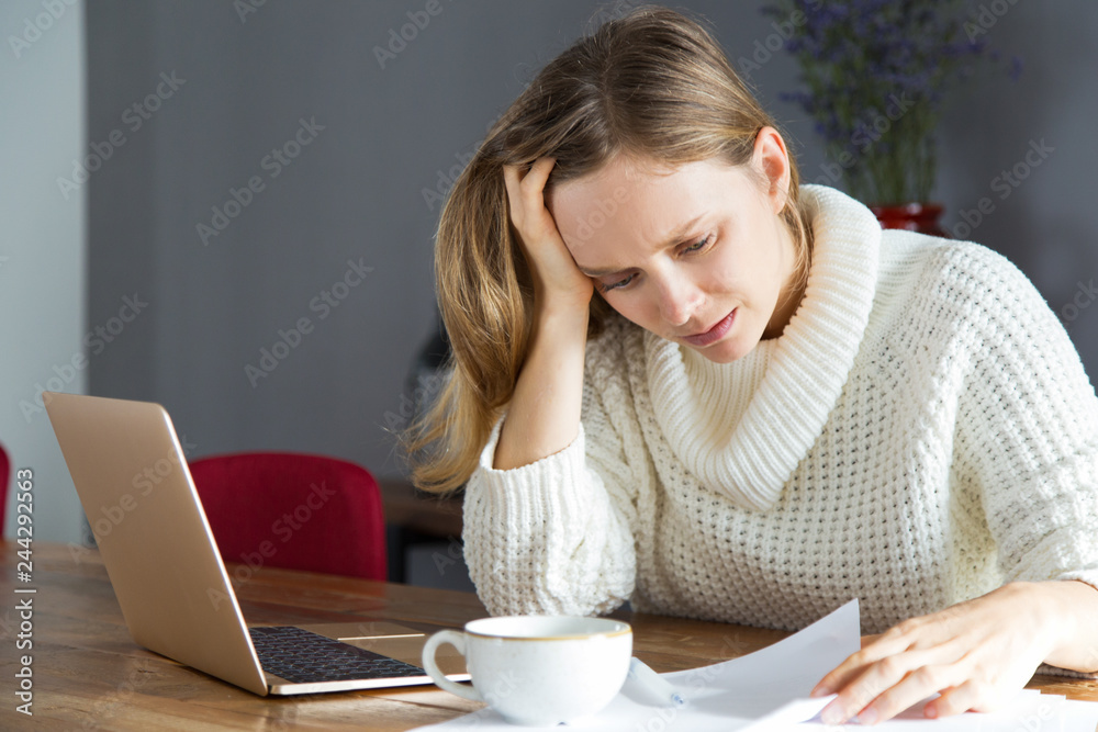 Fototapeta premium Confused young woman reading document at workplace. Upset lady in white sweater analyzing data. Mistake in report concept