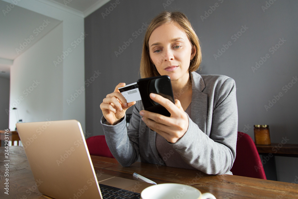 Young lady making online payment using smartphone. Pensive modern woman sitting at table and adding credit card number for mobile app. Online banking concept Young lady making online payment using smartphone. Pensive modern woman sitting at table and adding credit card number for mobile app. Online banking concept