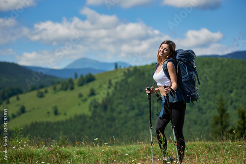 Smiling sporty female tourist with backpack and trekking poles, posing with closed eyes on sunny day. Mountains, forests and blue sky with clouds on the blurred background. Concept of active lifestyle