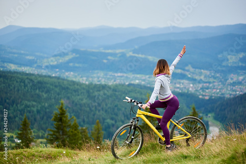 Sporty girl biker cycling on yellow mountain bike with her hand up on cloudy evening. Mountains, forests and small city on the blurred background. Outdoor sport activity, lifestyle concept