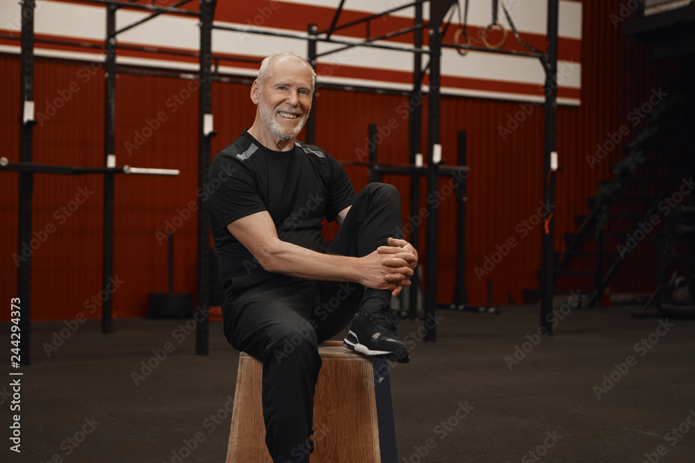 Portrait of happy healthy unshaven male pensioner in stylish sportswear relaxing on wooden chair in gym interior, smiling broadly at camera after crossfit workout with trainer. Fitness and activity Portrait of happy healthy unshaven male pensioner in stylish sportswear relaxing on wooden chair in gym interior, smiling broadly at camera after crossfit workout with trainer. Fitness and activity