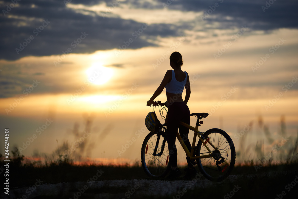 Obraz premium Back view of woman with bicycle standing on road among grass enjoying the sunset on evening sky. The girl has a beautiful sporty body. Helmet hangs on the handle of bicycle