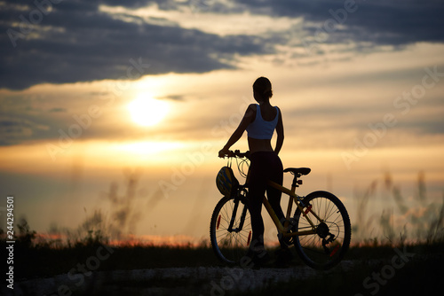 Back view of woman with bicycle standing on road among grass enjoying the sunset on evening sky. The girl has a beautiful sporty body. Helmet hangs on the handle of bicycle