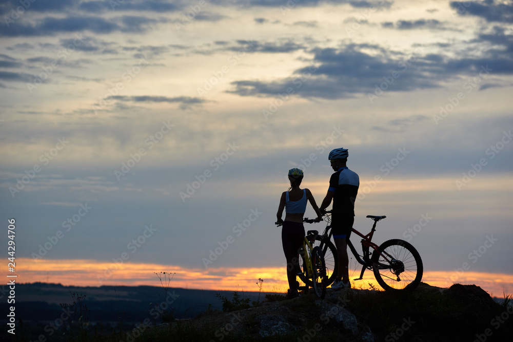 Rear view of two young people with mountain bikes stand on top of a cliff with beautiful scenery of hills and sky at sunset. The concept of a healthy lifestyle