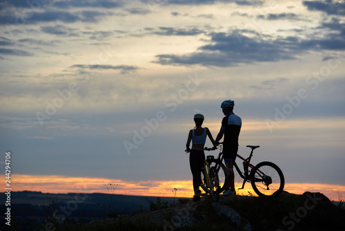 Fototapeta Naklejka Na Ścianę i Meble -  Rear view of two young people with mountain bikes stand on top of a cliff with beautiful scenery of hills and sky at sunset. The concept of a healthy lifestyle