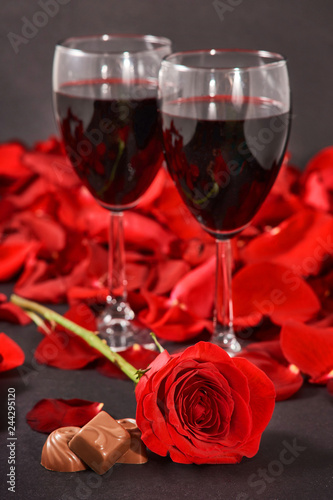 two glasses of wine, rose, petals and chocolates on a black background
