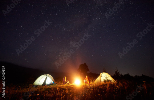 Night camping in mountains. Bright campfire burning between two tourists, boy and girl sitting opposite each other in front of illuminated tents under dark blue starry sky on distant hills background.